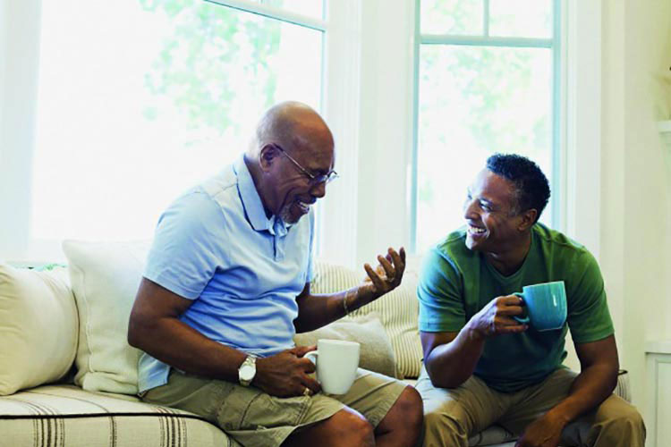 An older man and younger man sitting on a sofa in front of bay windows, having a conversation.